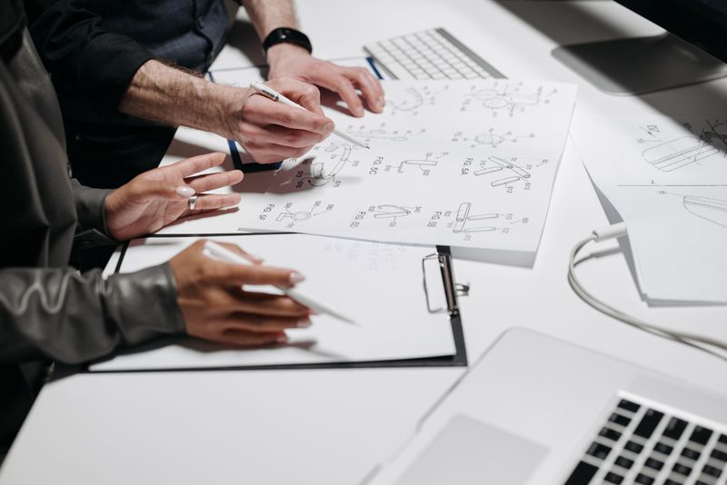 Two people reviewing printed technical drawings at a desk with laptops